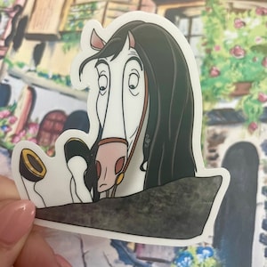 以下が含まれることがあります： 茶色の手綱をつけた、黒のたてがみと尻尾を持つ白い馬が、驚いた表情で横を向いている。馬は漫画風で、ステッカーの一部である。