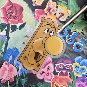 May include: A clear sticker with a cartoon image of a golden door knob with a face. The door knob has a large, round nose and a wide, open mouth. The sticker is on a background of colorful flowers.