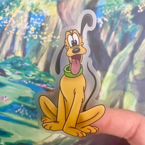 May include: A clear sticker featuring a cartoon image of Pluto, a yellow dog with black ears and a green collar. Pluto is sitting with his tongue sticking out and looking to the left.