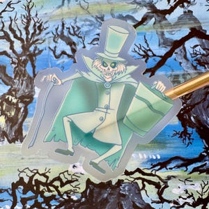 May include: A clear sticker featuring a cartoon ghost wearing a top hat and holding a bucket. The ghost is green and white and has a skeletal face. The background is a blurry image of trees.