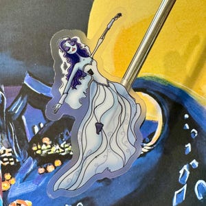 May include: A clear sticker featuring a cartoon illustration of a bride with long purple hair and a white wedding dress. The bride is in a dancing pose with one arm raised above her head.