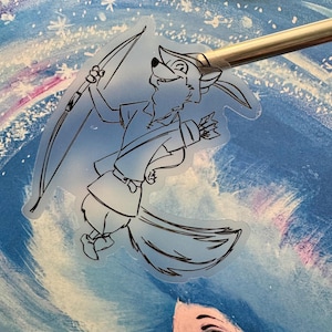 Op de afbeelding: Een transparante sticker met een cartoonvos, gekleed als Robin Hood, met een pijl en boog. De vos staat in een dynamische pose tegen een lichtblauwe achtergrond. De sticker wordt vastgehouden door een metalen tool.