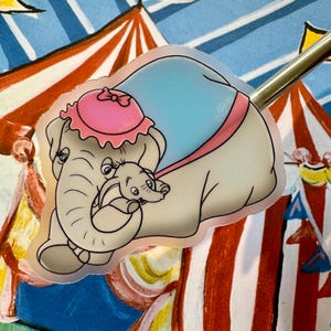 以下が含まれることがあります： ピンクとブルーのステッカーに、赤ちゃんゾウを抱っこした漫画のゾウが描かれています。ゾウは、リボンが付いたピンクの帽子をかぶっています。ステッカーは、赤と白のストライプの背景に貼られています。