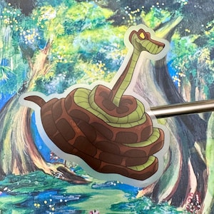 以下が含まれることがあります： 茶色の体と緑色のコイルを持つ漫画のヘビが描かれた透明なステッカー。ヘビは黄色い目と赤い舌をしています。ステッカーは、木と水のあるカラフルな塗装された背景に配置されています。