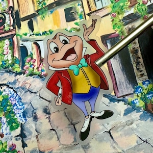 May include: A cartoon frog wearing a red jacket, yellow vest, blue pants, and a turquoise bow tie. The frog is smiling and has a surprised expression on its face. The frog is standing on a cobblestone street with a painted background of a building and flowers.
