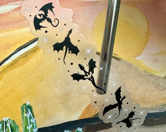 May include: A clear sticker featuring a design of four black silhouettes of dragons flying in a row against a white background with silver glitter accents.