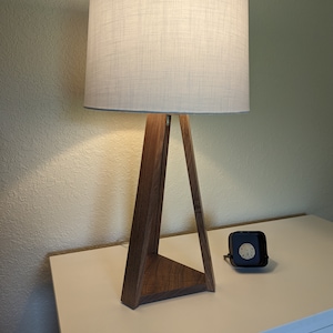 May include: A wooden table lamp with a white fabric shade. The lamp has a triangular base and is sitting on a white surface.