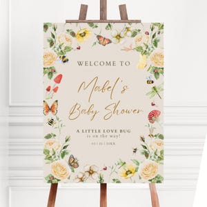 May include: A baby shower welcome sign on a wooden easel. The sign features the text "Welcome to Mabel's Baby Shower" in gold script, surrounded by a floral and insect border. The text "A Little Love Bug is on the way!" is also included.