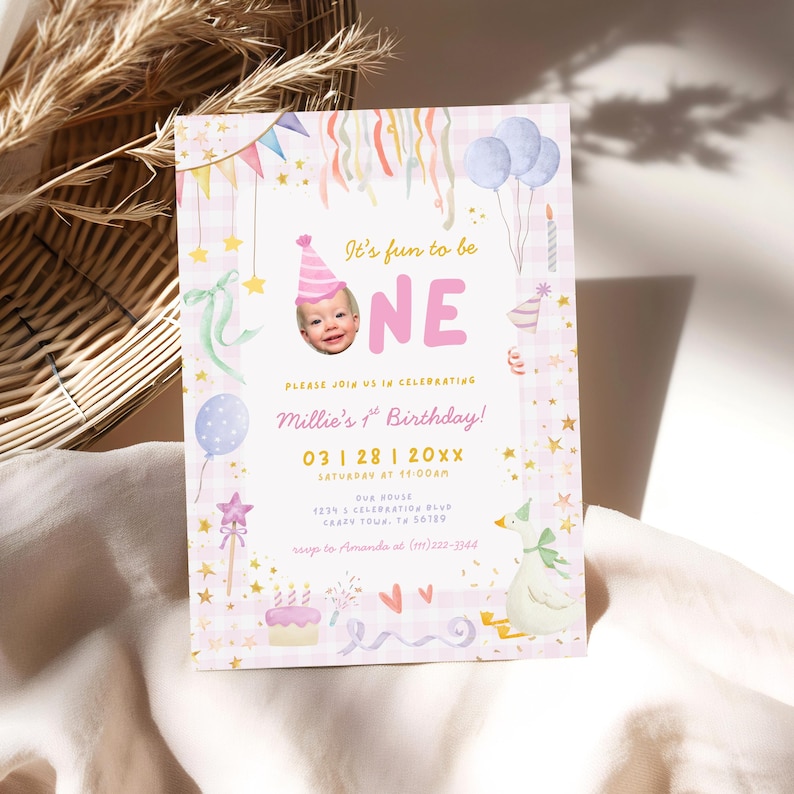 May include: A pink and white birthday invitation with the text "It's fun to be ONE". The invitation features a photo of a baby, balloons, stars, and a cake. The invitation is for Millie's 1st birthday.