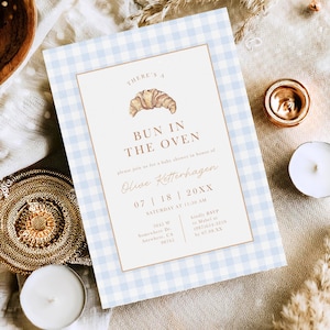 May include: Baby shower invitation with a light blue gingham border. The invitation features the text "There's a Bun in the Oven" with a croissant illustration. Details include the honoree's name, date, time, and RSVP information.