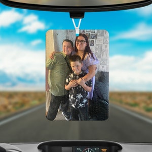 Peut inclure: Un désodorisant de voiture rectangulaire suspendu au rétroviseur, présentant une photographie imprimée de trois personnes ensemble. L'image montre une personne en chemise verte, une personne en haut violet clair et un enfant en chemise noire à palmiers.