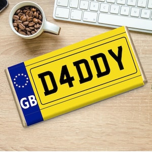 Op de afbeelding: Een gele reep met het woord "DADDY" in vette zwarte letters, die op een kentekenplaat lijkt. De reep heeft een blauwe streep met een cirkel van sterren en de letters "GB" aan de linkerkant. De reep ligt op een houten oppervlak.