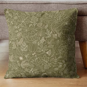 May include: Square cushion with a fall-themed leaf and pumpkin pattern. The cushion has a muted green background with white outlines of leaves, acorns, and pumpkins. The cushion is placed on a wooden floor next to a grey sofa.