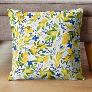 May include: Square throw pillow with a white background, featuring a pattern of yellow lemons, green leaves, and blue floral accents. The pillow is placed on a wooden floor next to a gray sofa.