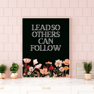 May include: Black and white typography print with the text "LEAD SO OTHERS CAN FOLLOW" on a black background with a floral border of pink, red, and orange flowers.