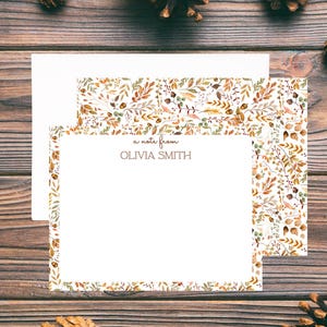 May include: Stationery set featuring a white notecard with the text "a note from OLIVIA SMITH" and a matching envelope. The stationery has a floral design with autumn colors. The background is a wooden surface with pinecones.