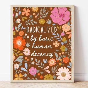 May include: A framed art print with a brown background and a floral design. The print features the words "RADICALIZED by basic human decency" in white script. The flowers are in shades of pink, orange, and yellow.