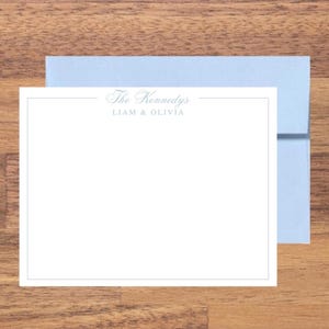 May include: A white stationery card with a light blue border and text that reads "The Kennedys" and "Liam & Olivia" in a script font. A light blue envelope is behind the card, set on a wooden surface.