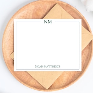 May include: A white stationery card with a dark blue border and the initials "NM" at the top. The name "NOAH MATTHEWS" is printed at the bottom. The card rests on a wooden plate with a brown envelope.
