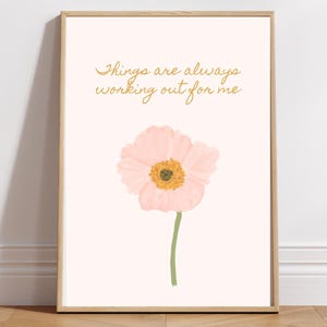 May include: A framed art print featuring a pink flower with a yellow center and a green stem. The artwork includes the gold text, "Things are always working out for me" on a light pink background. The frame is made of light wood.