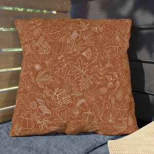 May include: A brown throw pillow with a white outline of autumn leaves, acorns, and pumpkins. The pillow is on a blue couch with a brown sun hat.