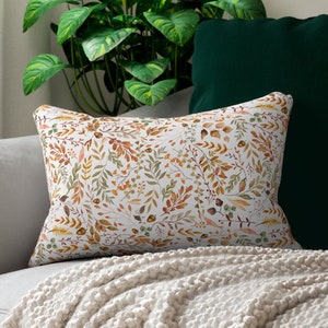 May include: A white cushion with a autumnal pattern of orange, brown and green leaves and branches. The cushion is on a white sofa with a green throw blanket.