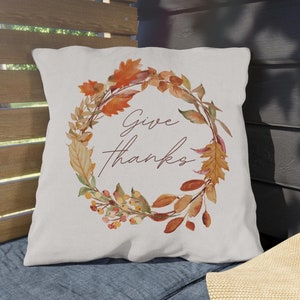 May include: A square, off-white decorative pillow with a fall-themed wreath design. The wreath is composed of orange, brown, and yellow leaves and berries. The words "Give thanks" are written in a cursive font within the wreath.