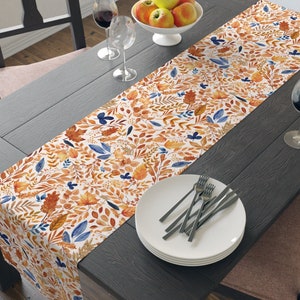 May include: A white table runner with a fall-themed design featuring orange, yellow, and blue leaves. The runner is laid on a dark wood table with a plate and silverware.