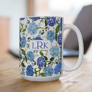Op de afbeelding: Witte keramische mok met een blauw hortensia bloemenpatroon. Het ontwerp bevat blauwe en lichtblauwe bloemen met groene bladeren en de initialen "LRK" in een witte rechthoek. De mok heeft een wit handvat.