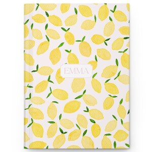 May include: A hardcover journal with a white background and a pattern of yellow lemons with green stems. The journal has the name "EMMA" printed on a pink label. The journal is a perfect gift for anyone who loves lemons.