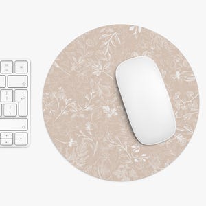 Könnte beinhalten: Ein runder, beigefarbener Mauspads mit einem weißen Blumenmuster, einer weißen Computermaus und einer weißen Tastatur. Das Mauspads befindet sich auf einer weißen Oberfläche. Das florale Design umfasst Blätter und Blüten.