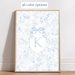 May include: A framed print with a light blue floral design. A light blue shield with a bow and the letter "K" is in the centre. The text "36 color options" is at the top. The frame is a light wood colour.