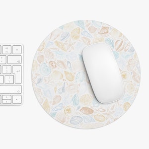 Puede incluir: Una alfombrilla de ratón redonda con fondo blanco y un patrón de conchas en tonos azules, rosas y amarillos. Un ratón de ordenador blanco está encima de la alfombrilla de ratón.