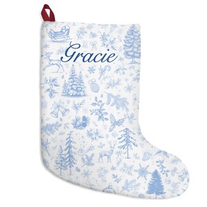 Puede incluir: Calcetín navideño blanco con un estampado azul toile de Jouy con árboles, ciervos y copos de nieve. El nombre "Gracie" está impreso en azul. Un lazo rojo está unido en la parte superior para colgar.