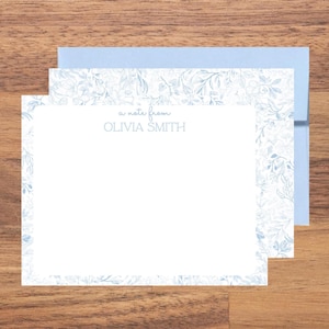 May include: A white rectangular stationery card with a light blue botanical leaf pattern border. The text "a note from" is printed in small blue script above the name "OLIVIA SMITH" in blue capital letters. A light blue envelope is behind.