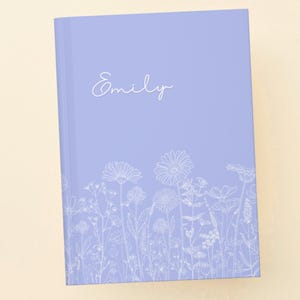May include: A lavender-colored journal with the name "Emily" in cursive script. The bottom of the journal features a white floral design. The journal is rectangular and appears to be closed.