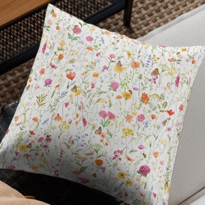 May include: A white throw pillow with a floral pattern featuring a variety of colorful flowers and leaves. The pillow is on a white couch with a brown wooden frame.