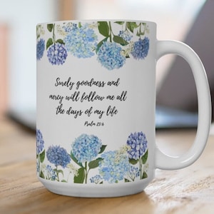 Op de afbeelding: Witte keramische mok met een bloemenontwerp met blauwe hortensia's. De mok heeft een citaat uit Psalm 23:6: "Zekerlijk zullen goedheid en genade mij volgen al de dagen van mijn leven."