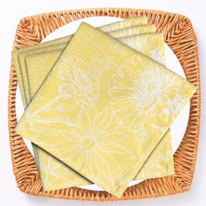 May include: A square wicker tray holds a white plate with four yellow napkins. The napkins feature a white floral design. A silver fork and knife are on either side of the tray, set against a white background.