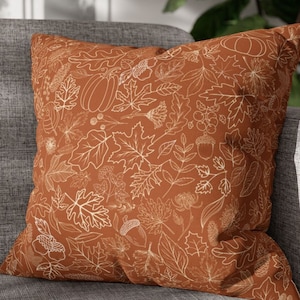 May include: A brown throw pillow with a white outline of fall leaves, pumpkins, and acorns. The pillow is on a gray couch.