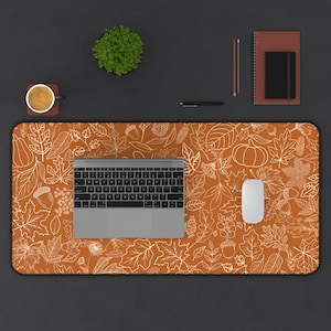 May include: A brown desk mat with a white outline of autumn leaves, acorns, and pumpkins. A laptop, a wireless mouse, and a cup of coffee are on the mat.