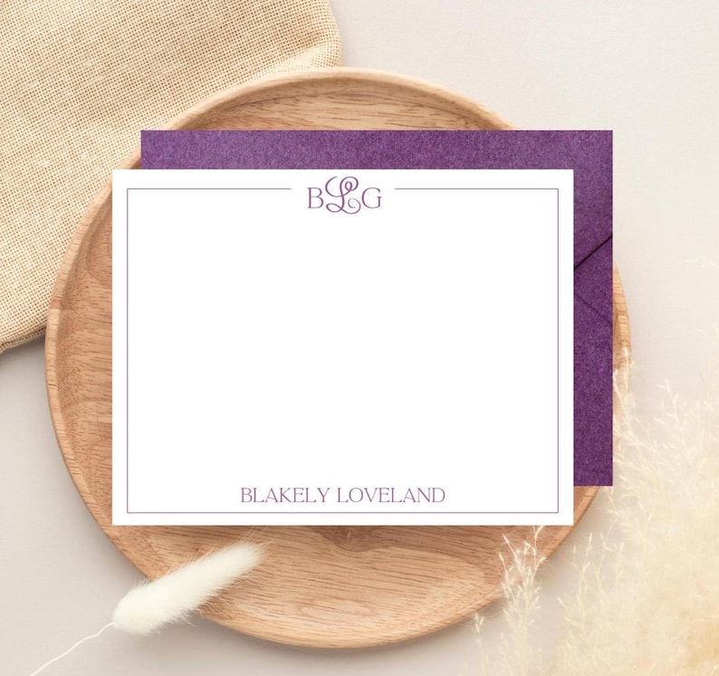 以下が含まれることがあります： 紫色のモノグラムと「Blakely Loveland」の名前が紫で印刷された白い便箋。カードは木製のプレートに置かれ、その背後には紫色の封筒があります。背景にはベージュの布と乾燥した植物が含まれています。