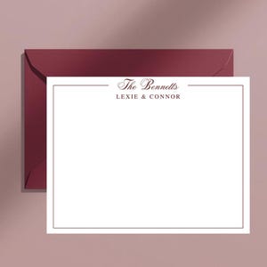 May include: A white stationery card with a burgundy border and the text "The Bennetts Lexie & Connor" in a script font. The card is on top of a burgundy envelope, with a soft pink background.