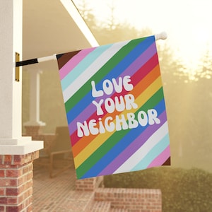 May include: A rainbow striped flag with the text "LOVE YOUR NEIGHBOR" in white. The flag is hanging from a pole on a porch.