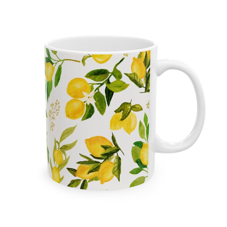 May include: White ceramic mug featuring a watercolor pattern of yellow lemons, green leaves, and white blossoms. The design is repeated around the mug's exterior. The mug has a white handle and is suitable for hot or cold beverages.