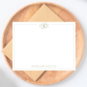 以下が含まれることがあります： 薄緑色のモノグラムと、お揃いのフォントで印刷された「Penelope Kelley」という名前が入った白い文房具セット。 文房具は、茶色の封筒が入った木製のプレートの上にあります。