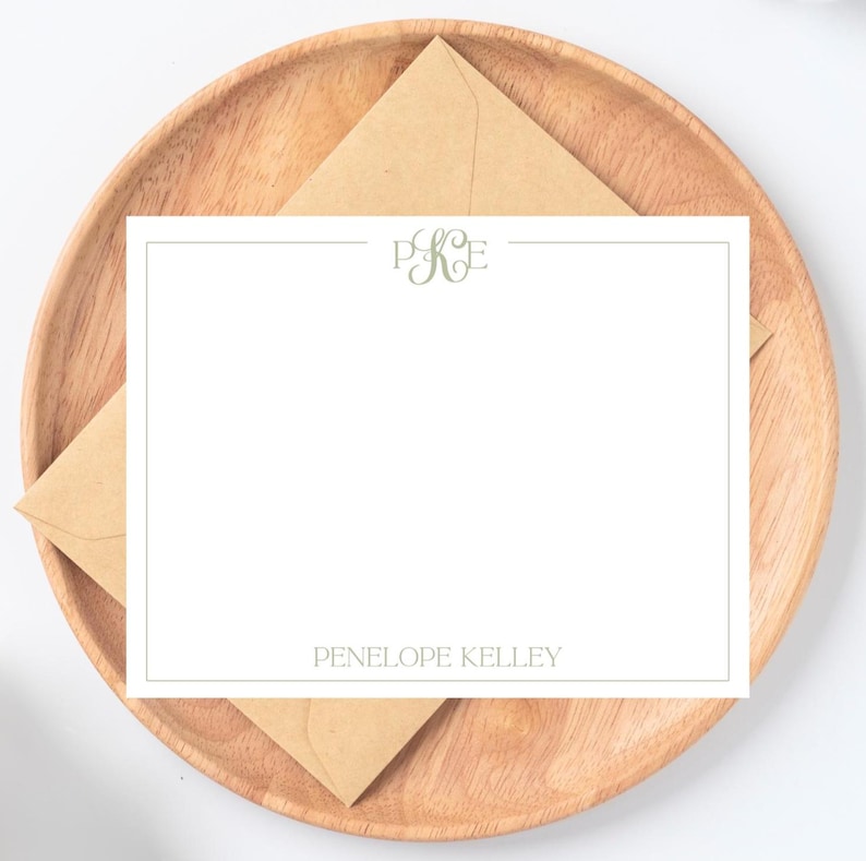 以下が含まれることがあります： 薄緑色のモノグラムと、お揃いのフォントで印刷された「Penelope Kelley」という名前が入った白い文房具セット。 文房具は、茶色の封筒が入った木製のプレートの上にあります。