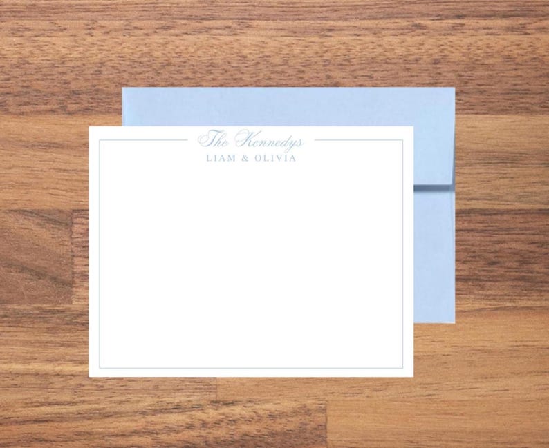 May include: A white stationery card with a light blue border and text that reads "The Kennedys" and "Liam & Olivia" in a script font. A light blue envelope is behind the card, set on a wooden surface.