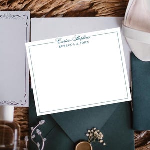 May include: A white stationery card with a teal border and the text "Carter-Hopkins REBECCA & JOHN" is centered on a dark teal envelope. The scene includes a white shoe, a decorative sprig, and a bottle.
