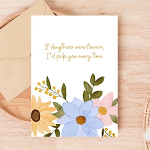 Peut inclure: Carte de v&oelig;ux blanche avec des fleurs &agrave; l'aquarelle jaunes, bleues et roses, et le texte "If daughters were flowers, I'd pick you every time." Une enveloppe en papier kraft est &agrave; c&ocirc;t&eacute; de la carte.